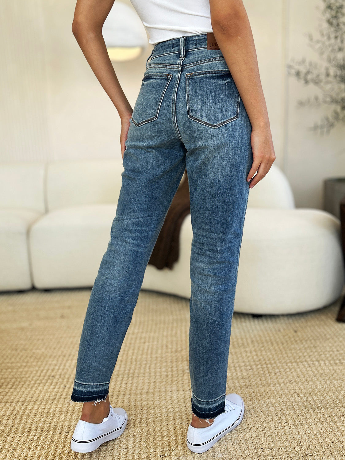 Person wearing a black top, Judy Blue Full Size Mid Rise Rigid Magic Release Hem Jeans, and black sneakers with white laces, standing on a beige carpet with hands in pockets.