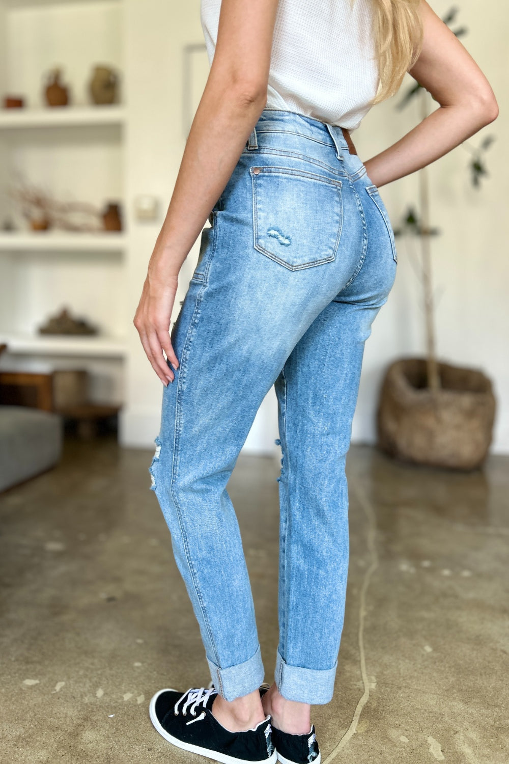 Two women are standing side by side, both clad in black and white tank tops and Judy Blue Full Size Distressed Straight Jeans with Patch Pockets, smiling at the camera in a modern living room.