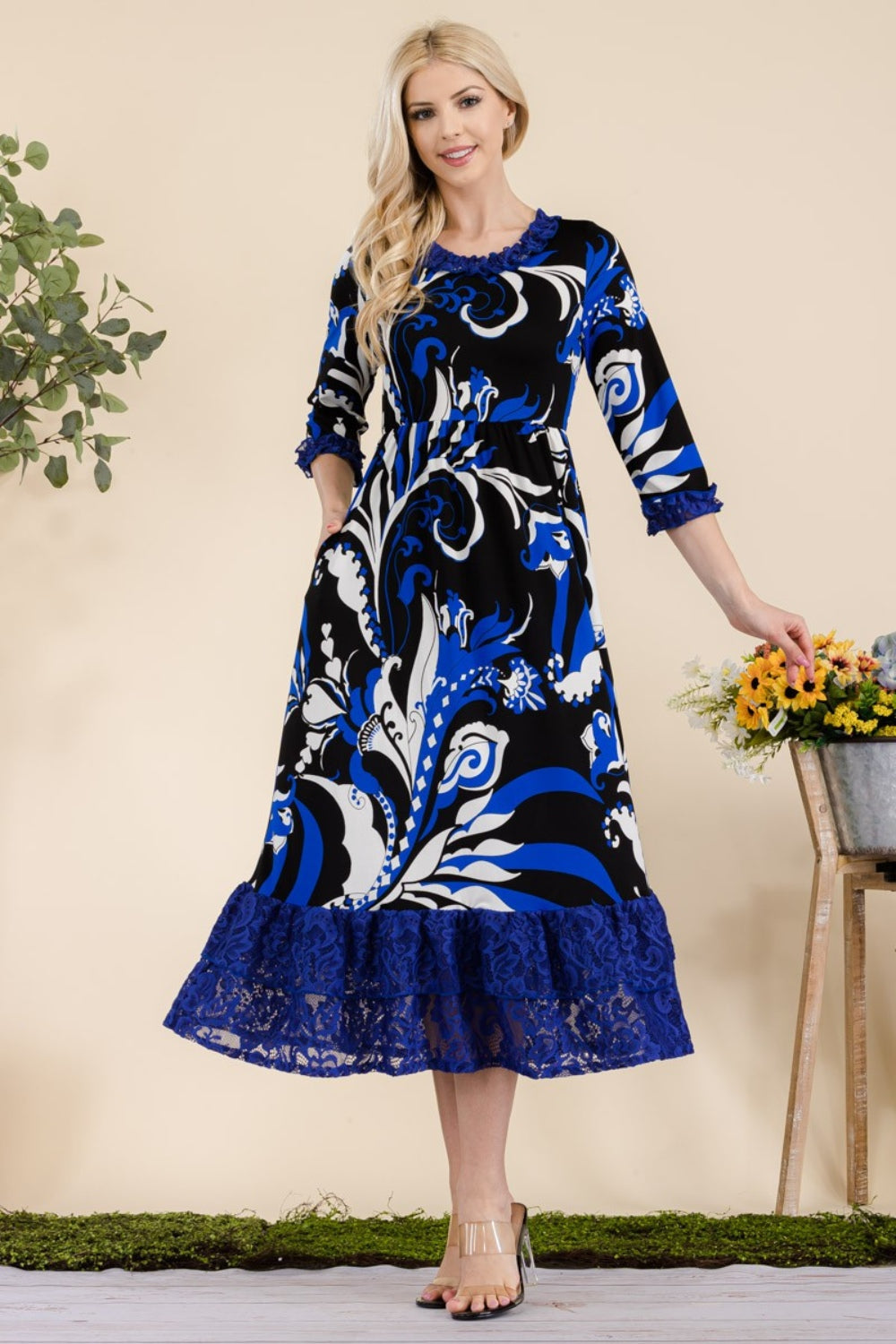 A woman wearing the Celeste Full Size Paisley Print Lace Ruffled Midi Dress stands near a wooden table adorned with a bucket of flowers.