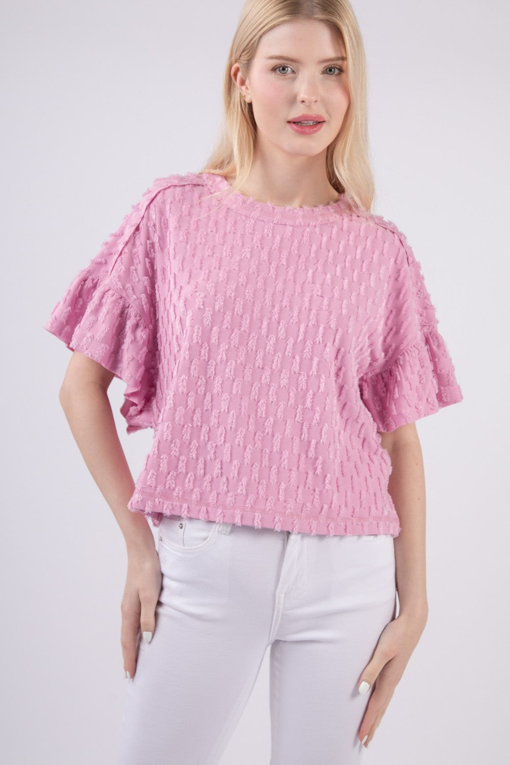 Person wearing the VERY J Full Size Texture Ruffle Short Sleeve Top, a chic and feminine textured pink blouse, paired with white pants, standing against a plain background.