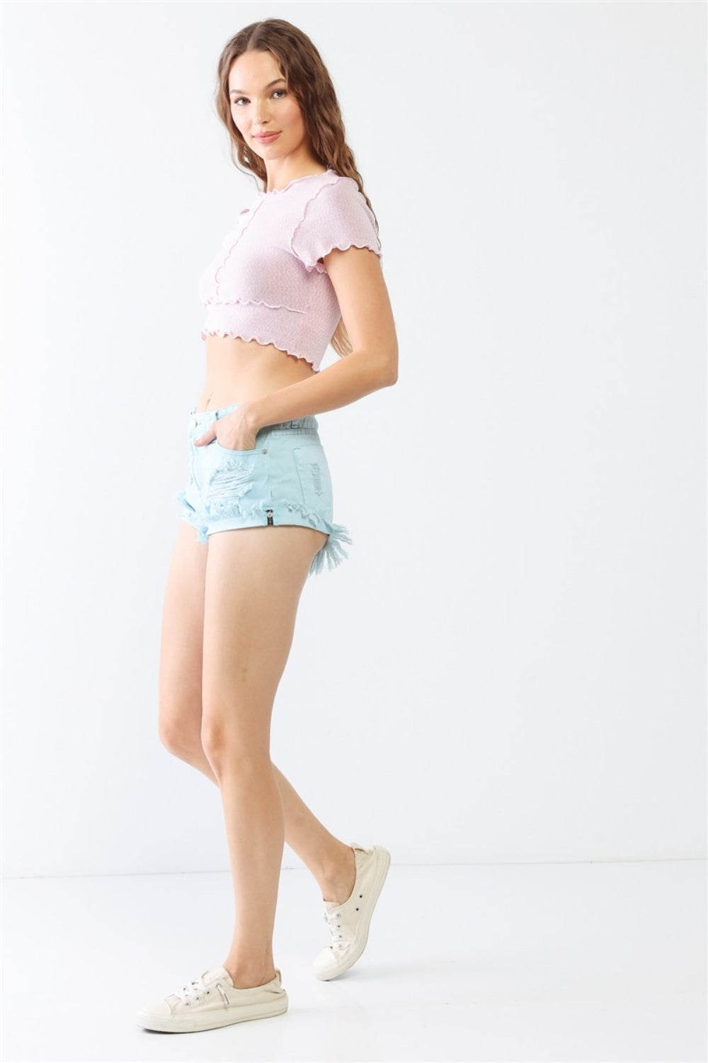 A person stands against a plain background, wearing a light pink crop top and LITZ LA Distressed Ripped Denim Shorts. They have long, wavy hair, embracing a trendy summer style.