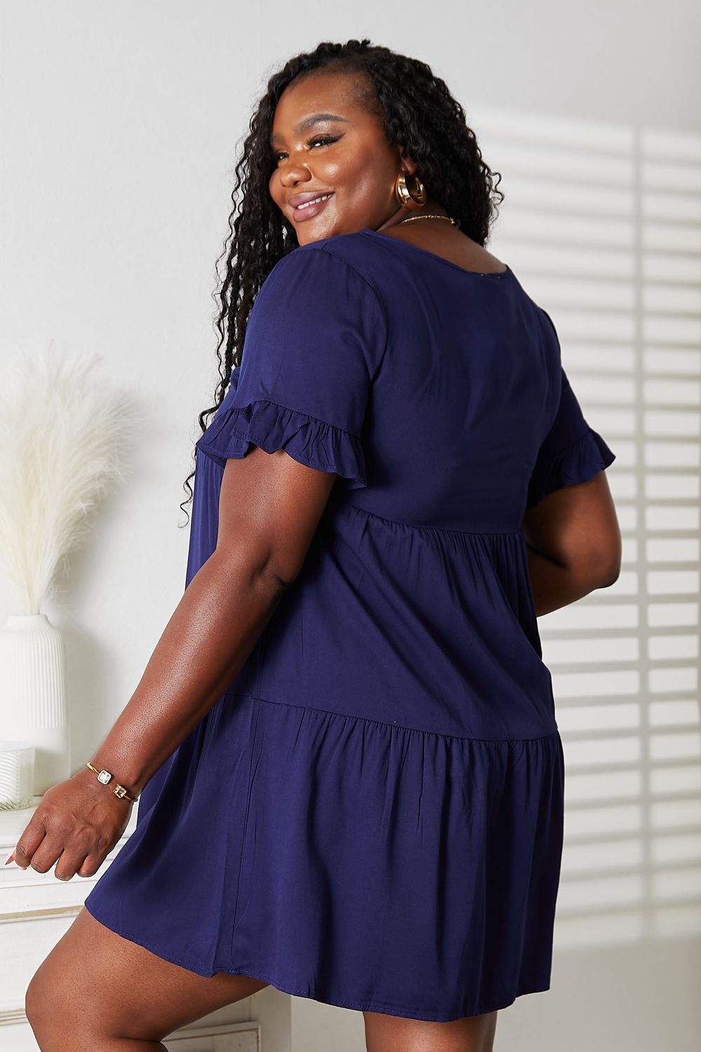 A woman stands indoors wearing the Mandy V-Neck Flounce Sleeve Tiered Dress in navy blue. She has one hand on her hip and is smiling.