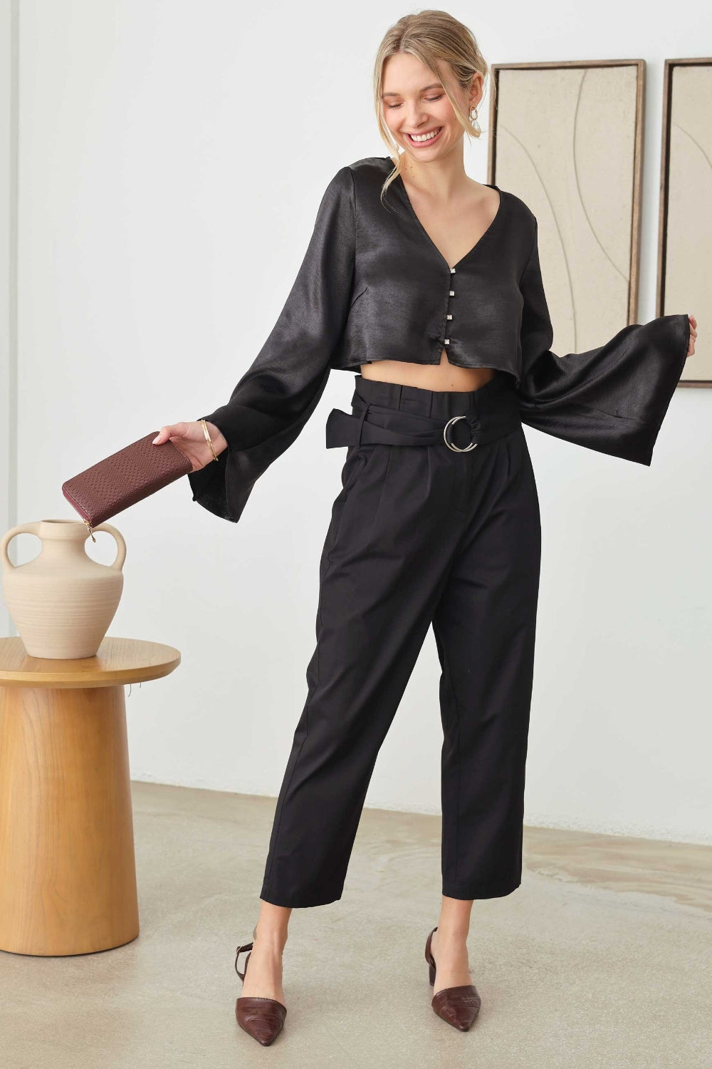 A person in a black long-sleeve Mustard Seed Satin Crop Top with rhinestone button closures and pants, holding a clutch, stands near a wooden table with a vase. Two framed artworks elegantly hang on the wall behind.