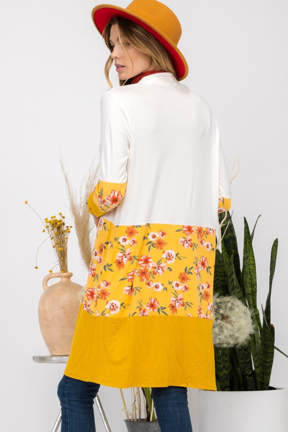 Wearing the Celeste Full Size Floral Color Block Open Front Cardigan with a white and yellow floral print, along with a red hat, a red top, and jeans, a woman stands beside a houseplant.