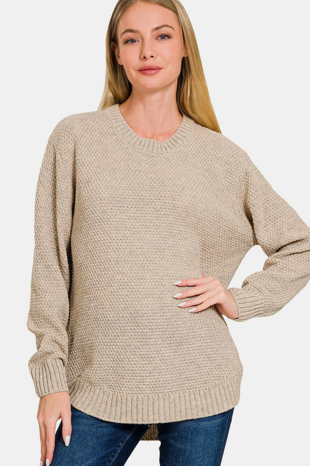 A person wearing a Zenana Full Size Round Neck Long Sleeve Curved Hem Sweater and jeans smiles while gently holding their neck.