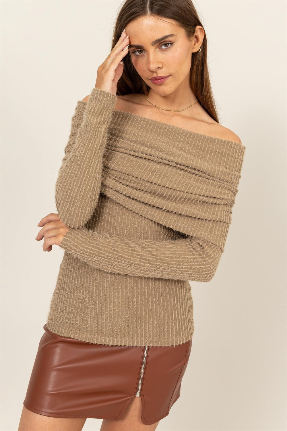 A woman elegantly stands against a light background, wearing the chic HYFVE Fuzzy Off Shoulder Textured Knit Top paired with a brown leather skirt.