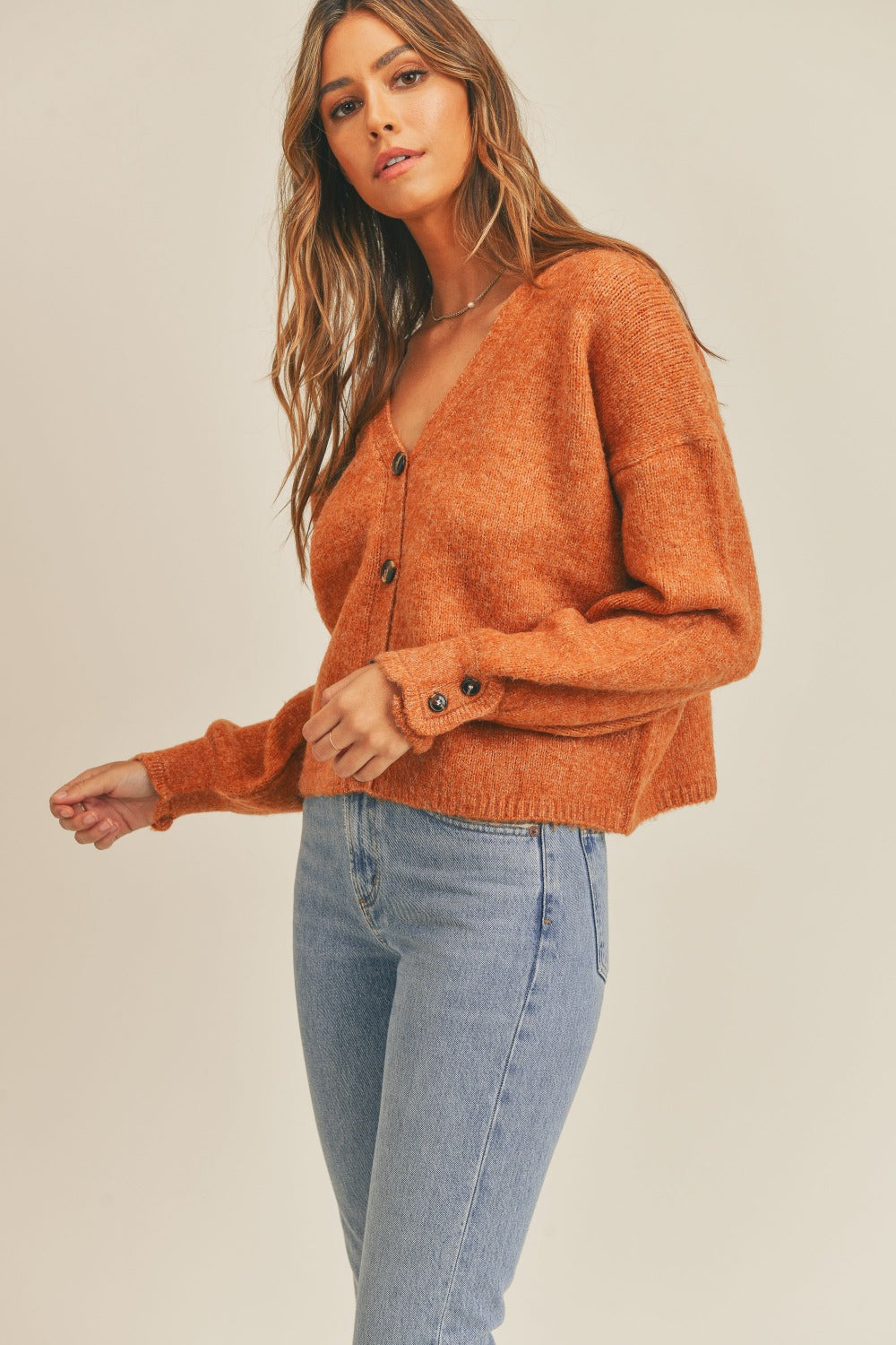 A woman wearing a MABLE Long Sleeve Button Down Sweater Cardigan in burnt orange and light blue jeans looks to her left against a plain background, exuding classic elegance.