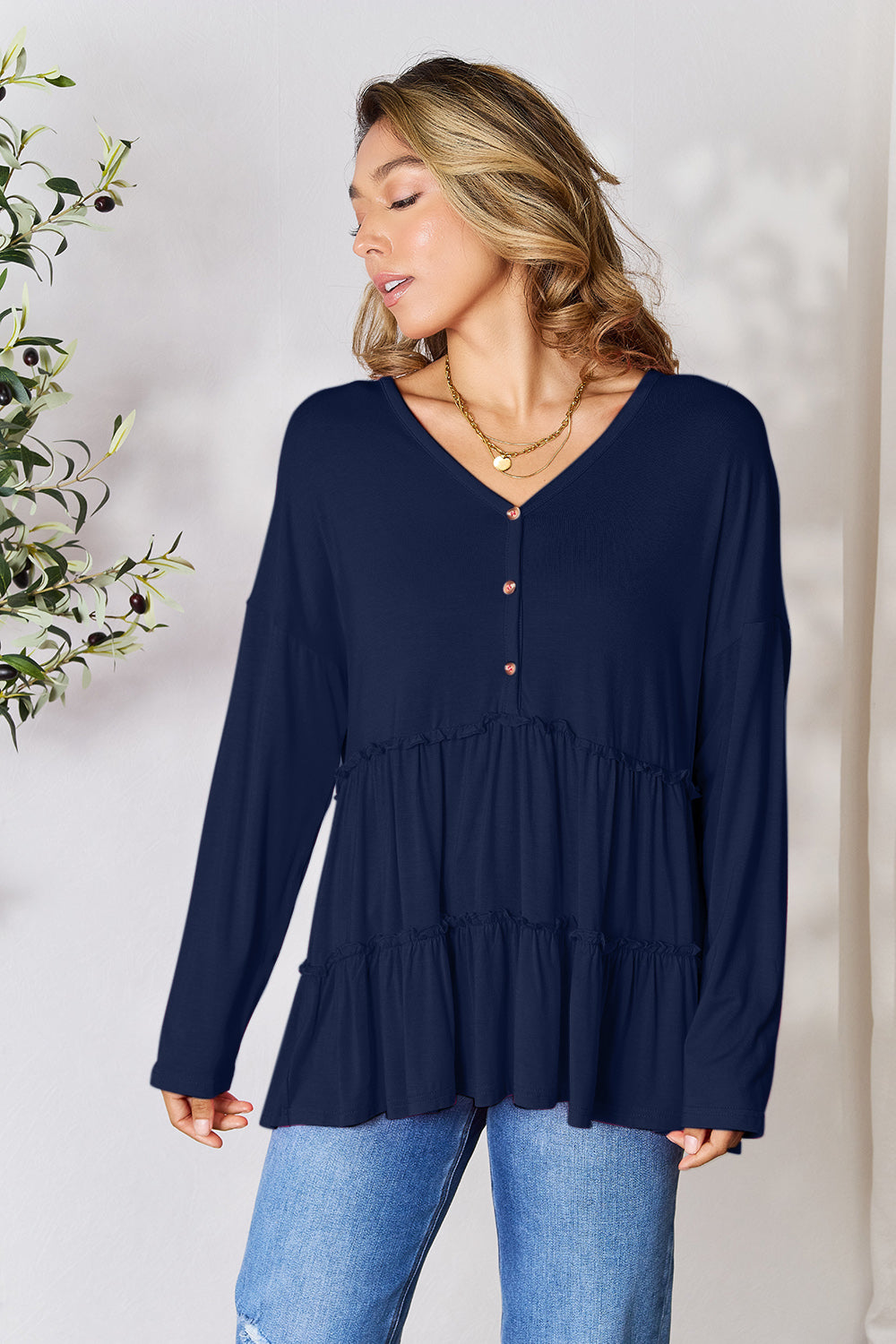 Person stands indoors near a plant, wearing the Double Take Half Button Long Sleeve Ruffle Hem Blouse in gray and blue jeans, smiling while looking to the side. The slightly stretchy fabric of the blouse adds comfort while its buttoned design offers a touch of elegance.