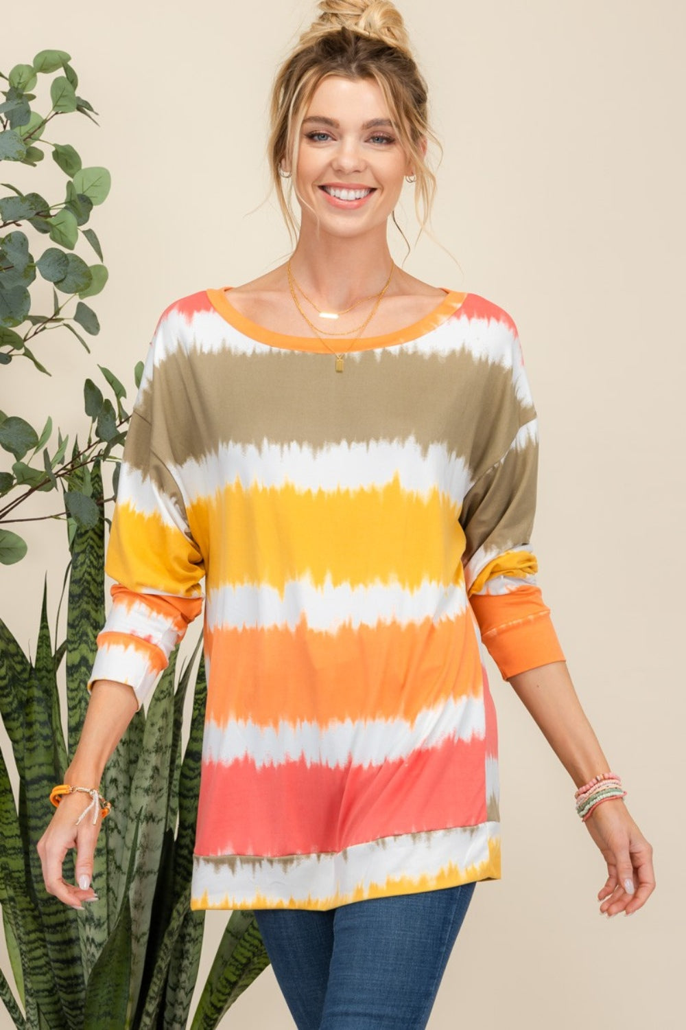 Wearing the Celeste Full Size Striped Long Sleeve T-Shirt paired with jeans, a person smiles while standing against a light background adorned with plants, exuding a casual yet stylish look.