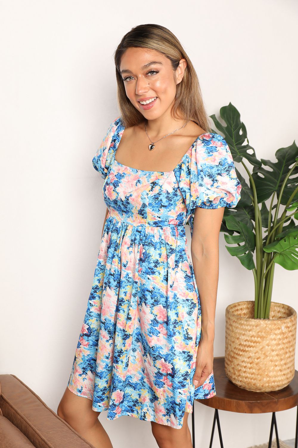 A woman in the Honey Floral Square Neck Puff Sleeve Dress stands smiling indoors, with a plant and a brown chair visible in the background.