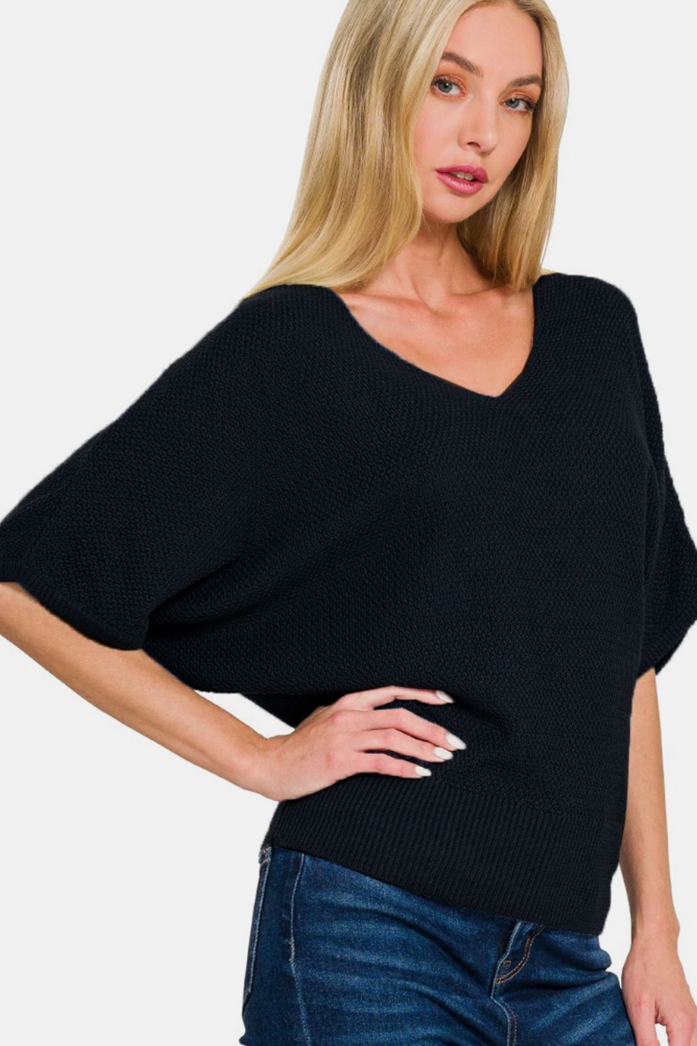 A person with long, blonde hair is wearing a Zenana V-Neck Short Sleeve Dolman Sweater in black and blue jeans, standing against a plain white background.