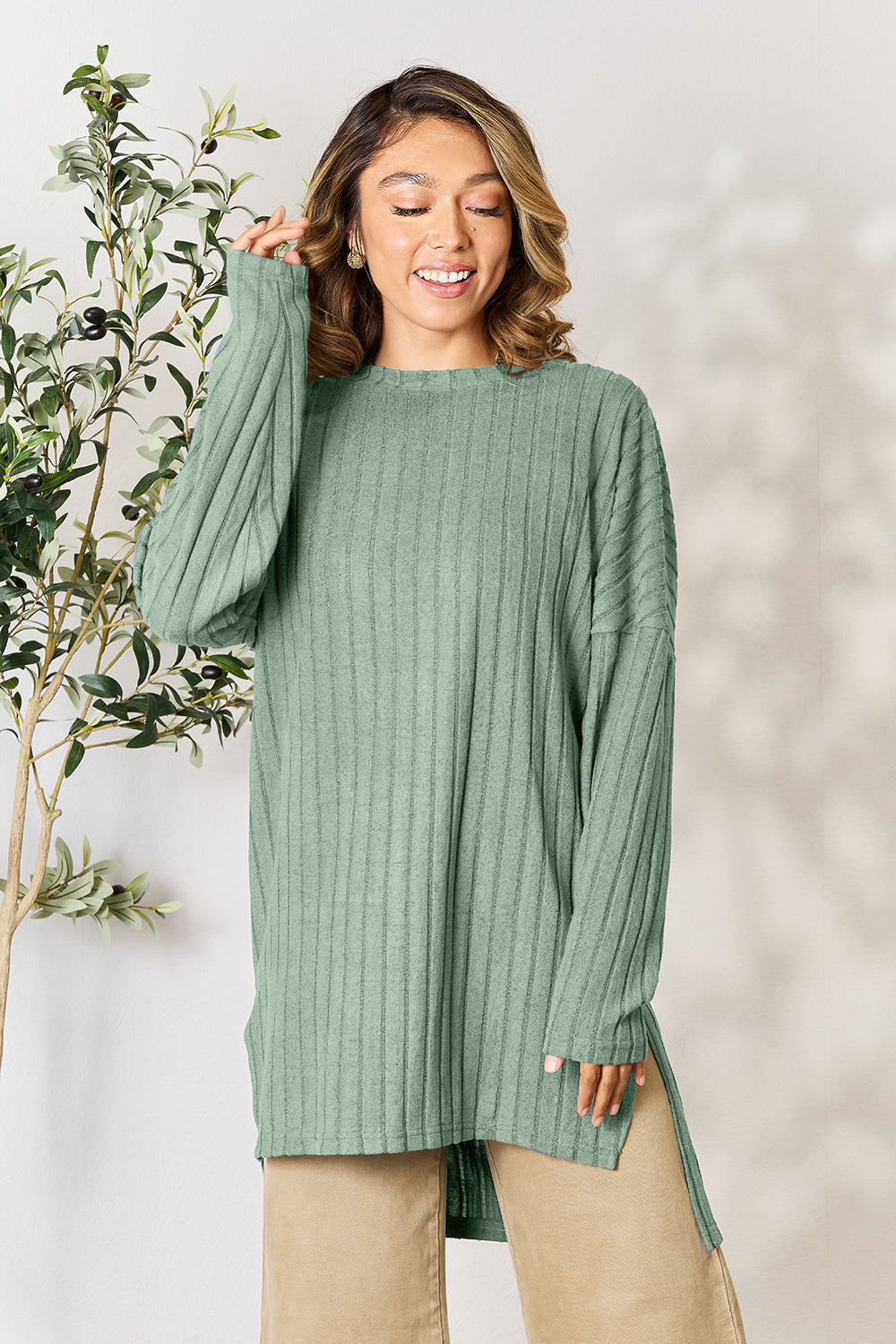 A person with shoulder-length hair smiles while wearing a beige ribbed sweater called the Basic Bae Full Size Ribbed Round Neck Long Sleeve Slit Top, along with matching beige pants, showcasing a basic style. They stand next to a green plant.