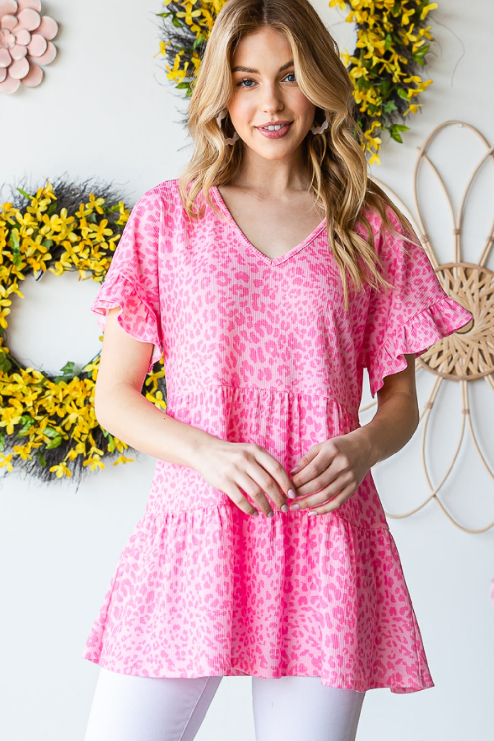 A person wearing a Heimish Full Size Leopard Ruffle Trim Short Sleeve Top stands in front of a floral wreath display.