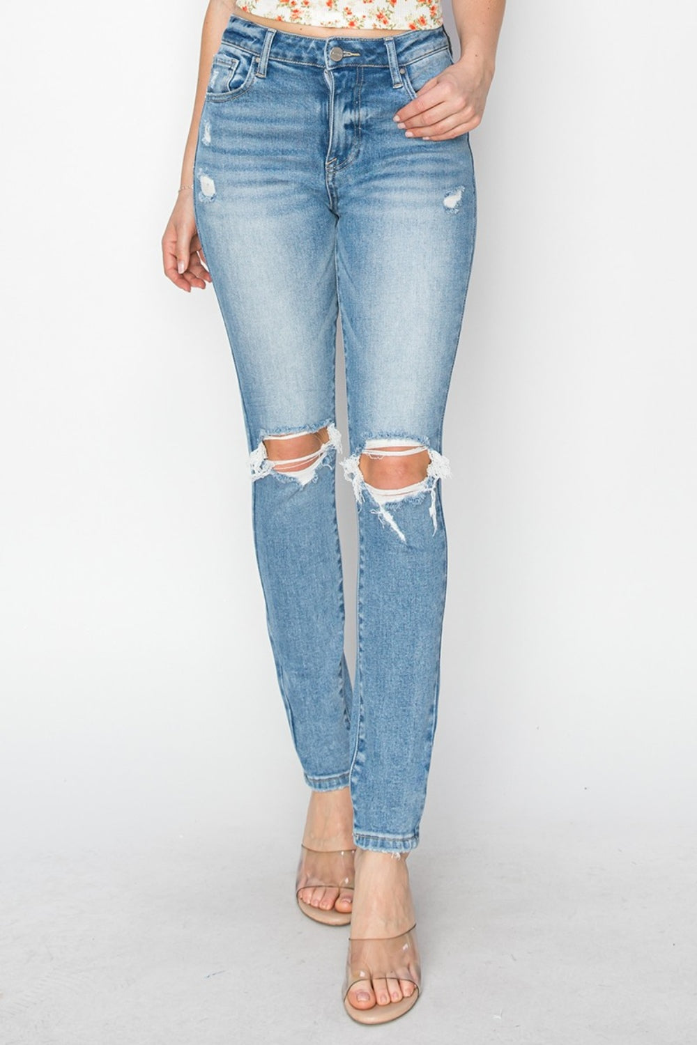 Wearing the Risen Full Size High Rise Knee Distressed Skinny Jeans in light blue, paired with a floral top and clear heeled sandals, showcases a fashion-forward wardrobe.