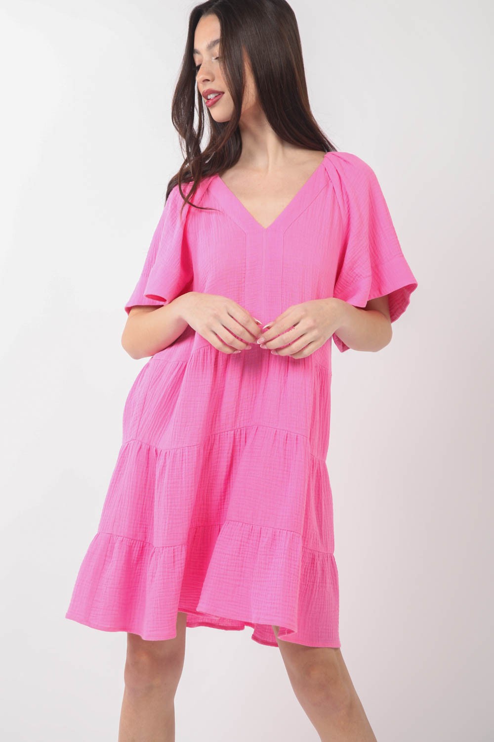 A woman is posing against a plain background, wearing the VERY J Texture V-Neck Ruffled Tiered Dress in pink, featuring short sleeves.