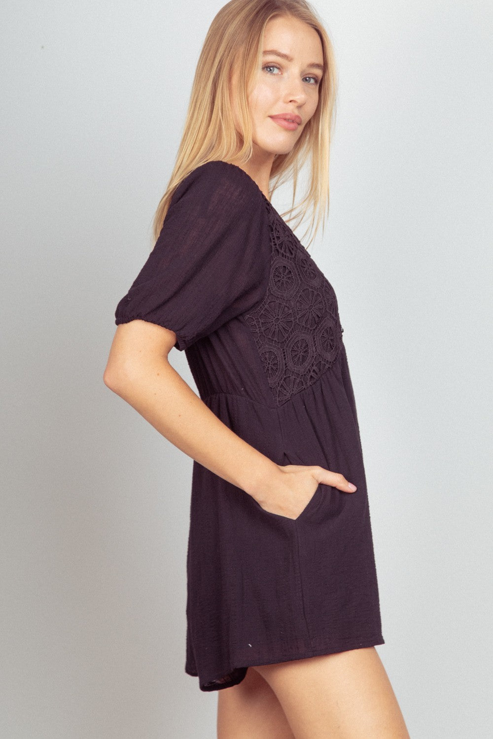 A woman with long blond hair stands against a neutral background, smiling with her hands in the pockets of her black VERY J Lace Detail Puff Sleeve Romper.