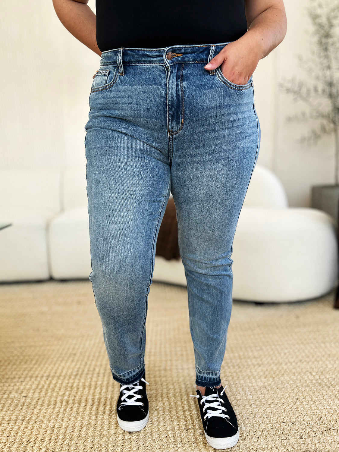 Person wearing a black top, Judy Blue Full Size Mid Rise Rigid Magic Release Hem Jeans, and black sneakers with white laces, standing on a beige carpet with hands in pockets.