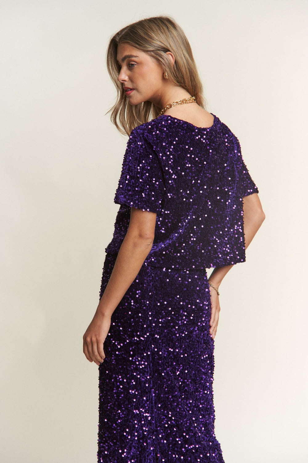 A woman wears a J.NNA Sequin Short Sleeve Crop T-Shirt in purple, with her eyes closed and arms raised above her head. She has long, wavy hair and is standing against a neutral background, creating a stylish addition to the scene.