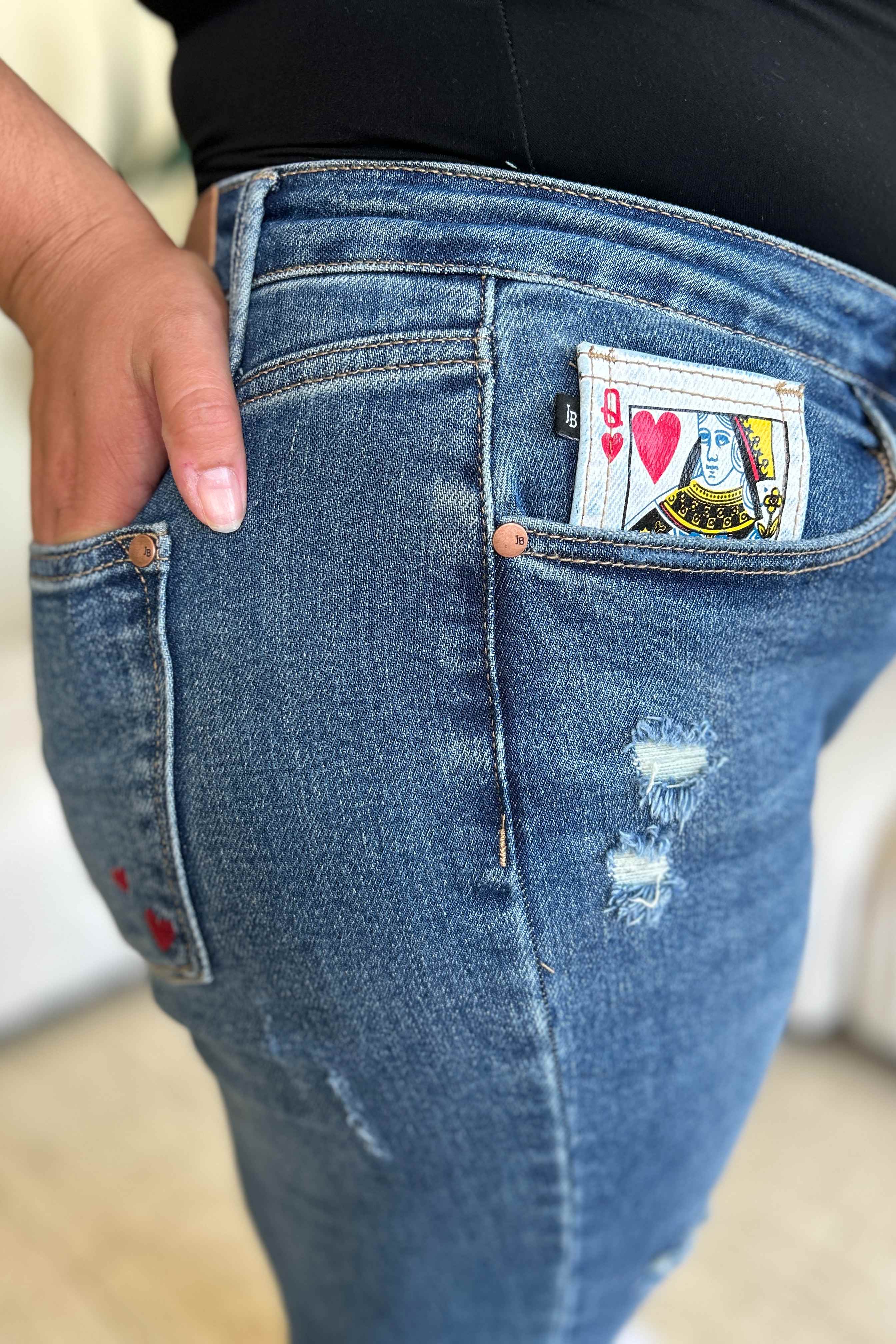 Two women stand side by side in a living room, both wearing stylish jeans and tank tops. One woman is pointing at the distinctive back pocket of her Judy Blue Full Size Queen Of Hearts Coin Pocket BF Jeans, while the other smiles.