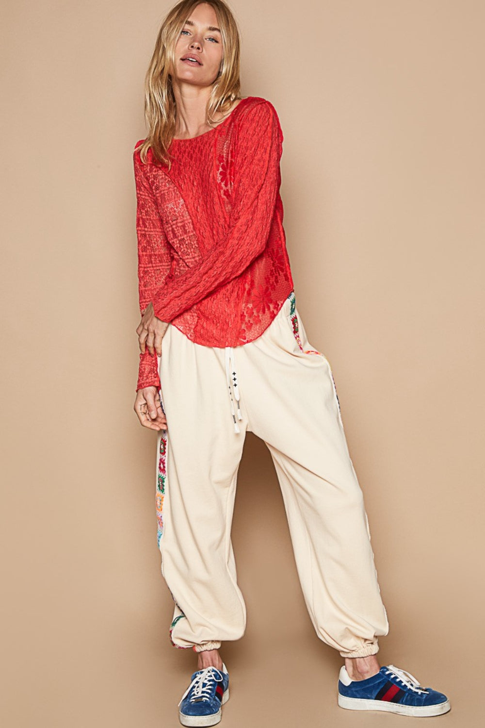 A woman with long blonde hair poses with one hand on her head, wearing a red POL Exposed Seam Long Sleeve Lace Knit Top and cream-colored patterned pants.