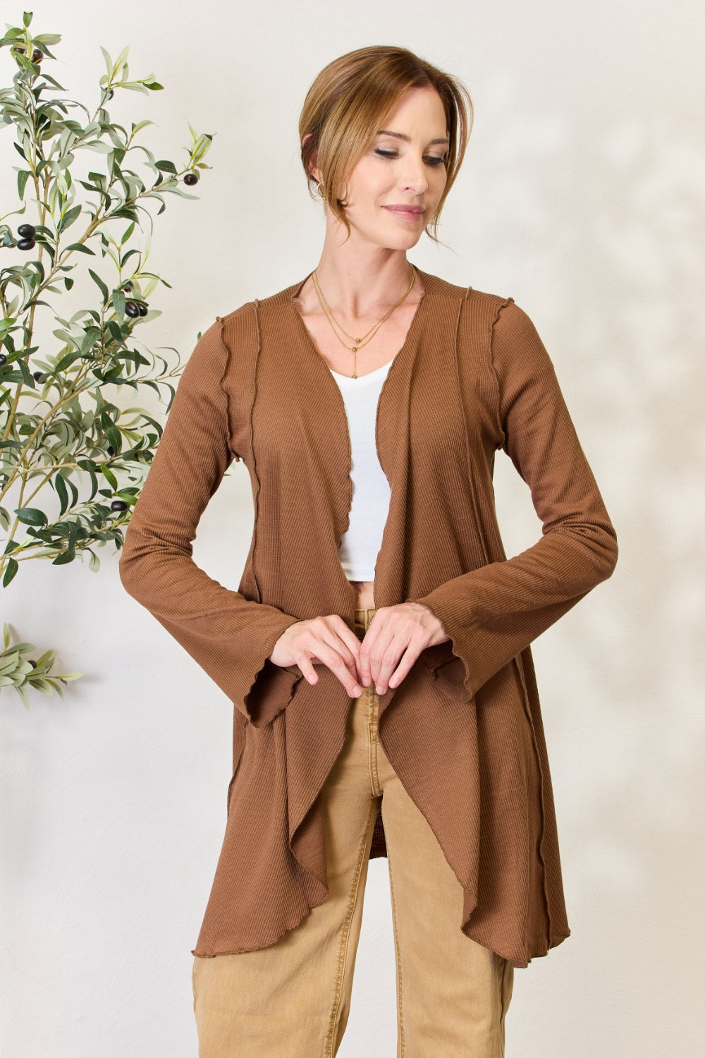 A woman stands in front of a light-colored wall and a small green plant, wearing the Culture Code Full Size Open Front Long Sleeve Cardigan with a merrow edge finish over a white top and beige pants.