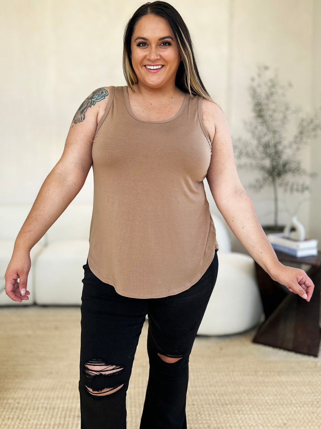 A woman wearing sunglasses, blue jeans, and the Basic Bae Full Size Round Neck Curved Hem Tank is standing in front of a concrete wall, holding her glasses with one hand and carrying a beige shoulder bag. Her basic style complements the slightly stretchy fabric of her outfit, ensuring comfort while maintaining chic simplicity.