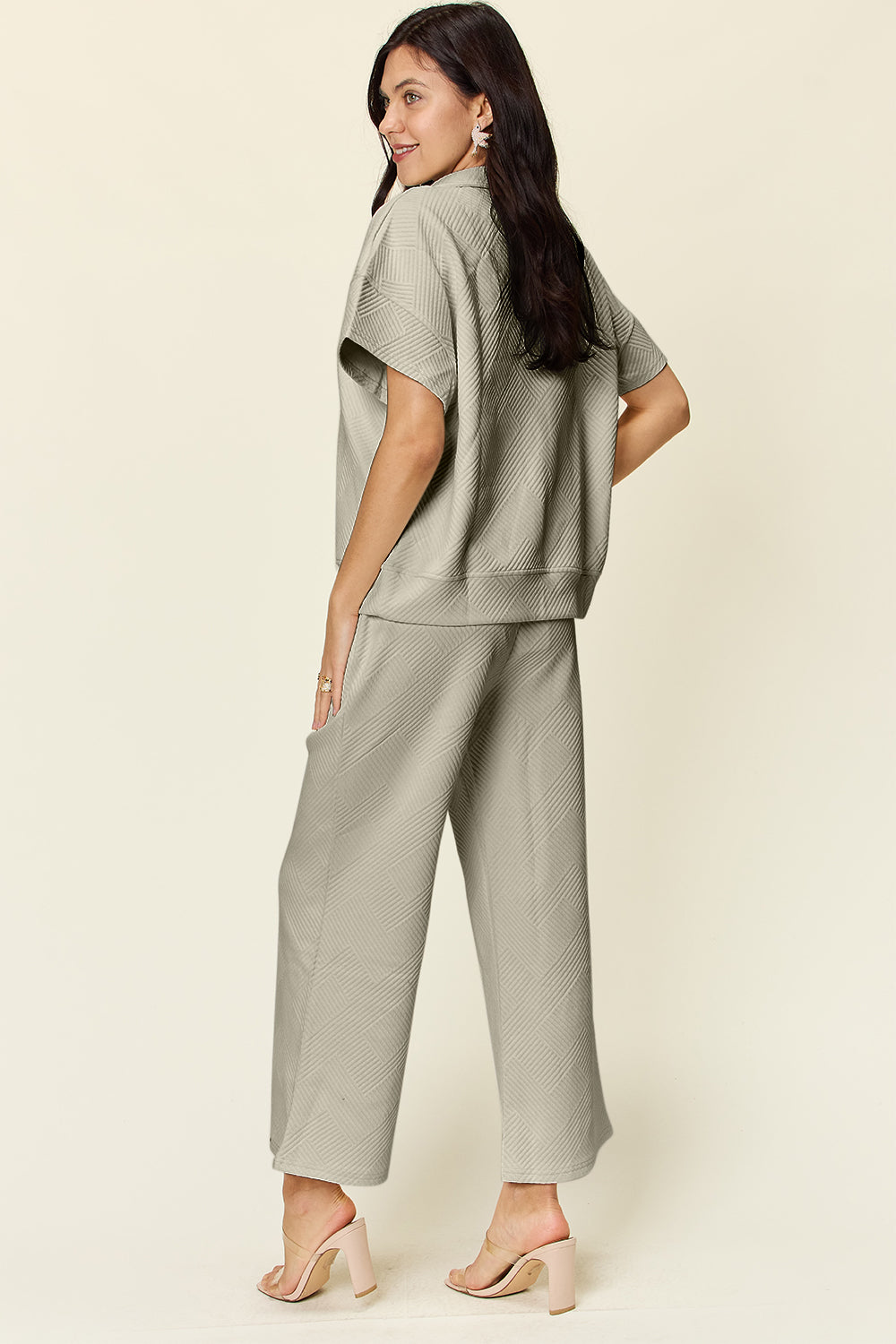 A woman is wearing the Double Take Full Size Texture Half Zip Short Sleeve Top and Pants Set, featuring a white zip-up short-sleeve top and matching wide-leg, drawstring pants. She stands with one hand in her pocket and smiles against a light background.