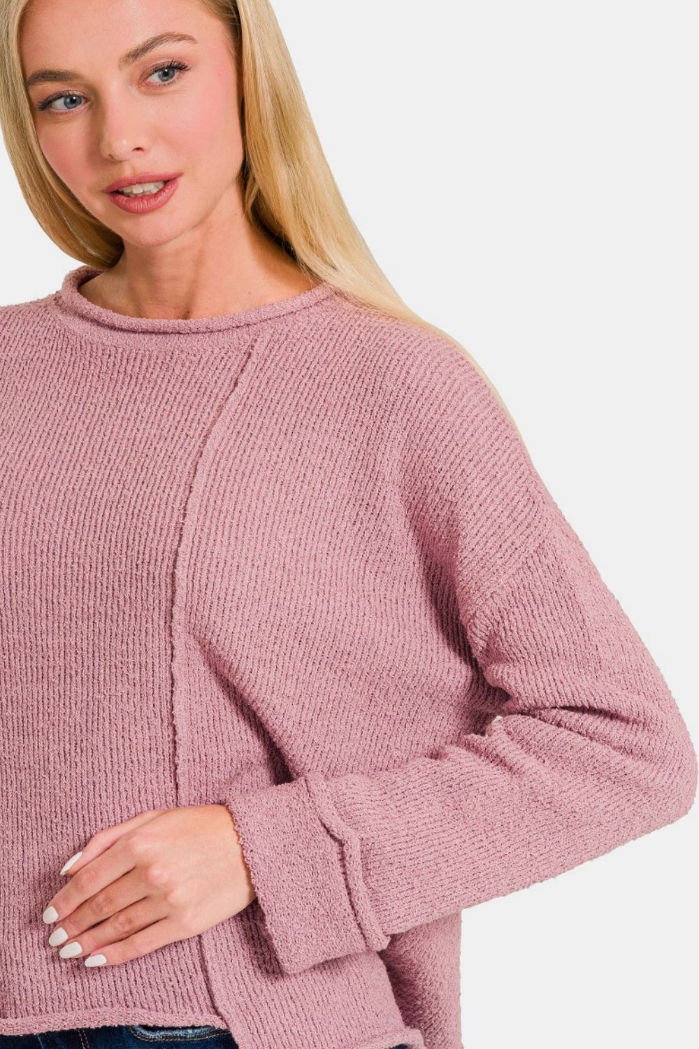 A person with long blonde hair is wearing a Zenana Asymmetric Hem Drop Shoulder Sweater in light pink and blue jeans, standing against a plain white background.