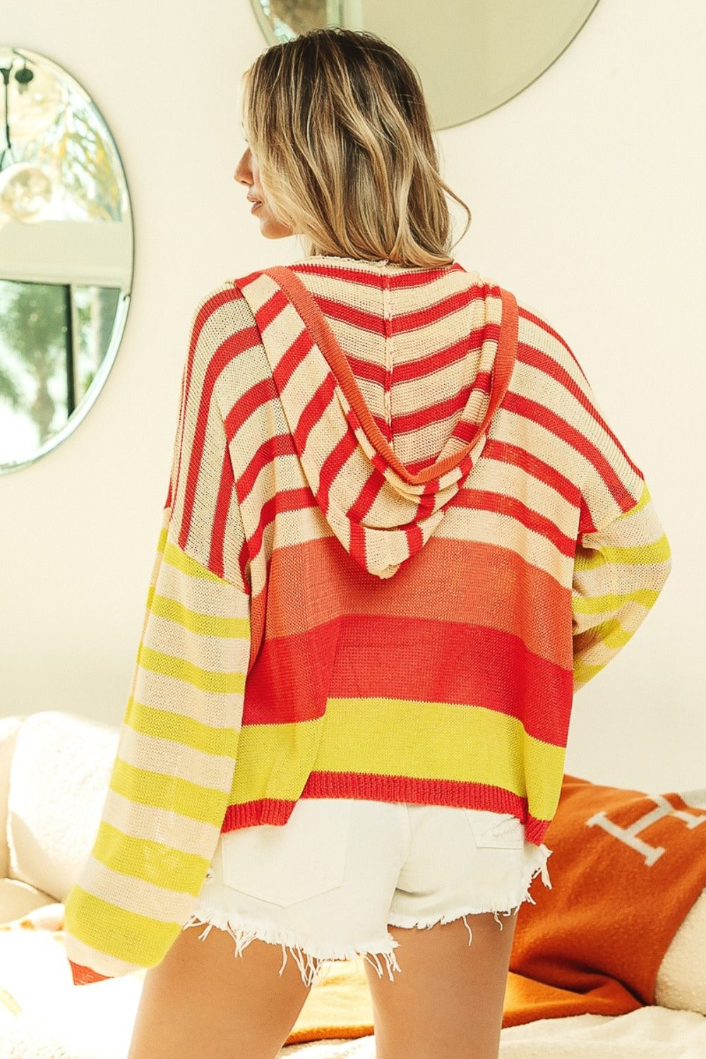 Person wearing a vibrant BiBi Striped Color Block Hooded Knit Top, paired with white shorts, standing in a bright room with round mirrors in the background.