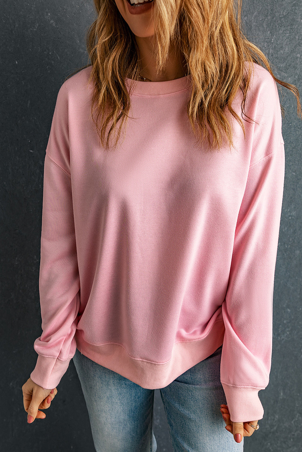 Individual with wavy brown hair wearing a Pink Plain Crew Neck Pullover Sweatshirt and blue jeans, standing with their back to the camera.