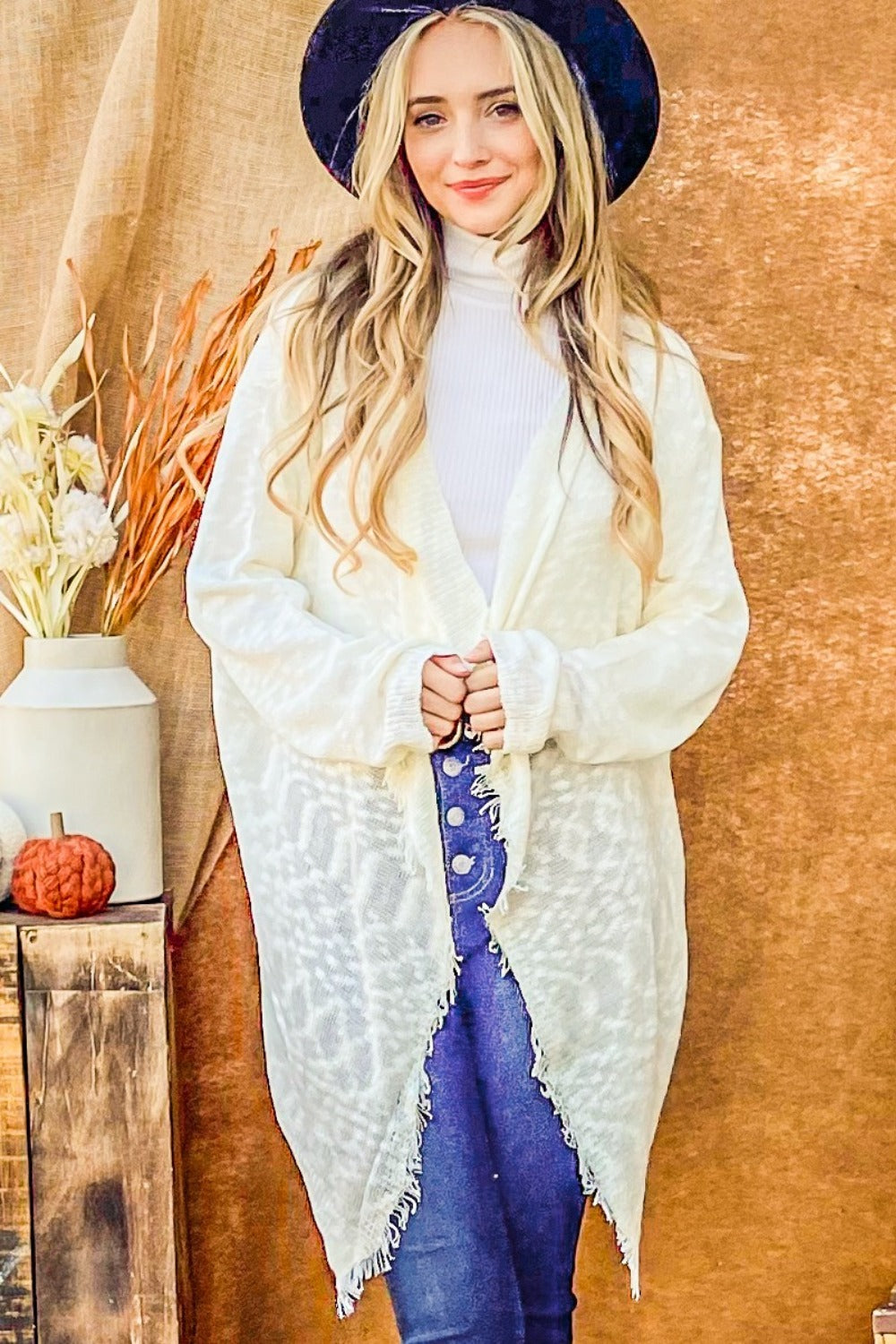 A person with long blonde hair wears a black hat, the And The Why Textured Knit Fringe Hem Cardigan in white, and blue jeans, standing against a brown backdrop near a wooden table with a vase of dried flowers and a small pumpkin.