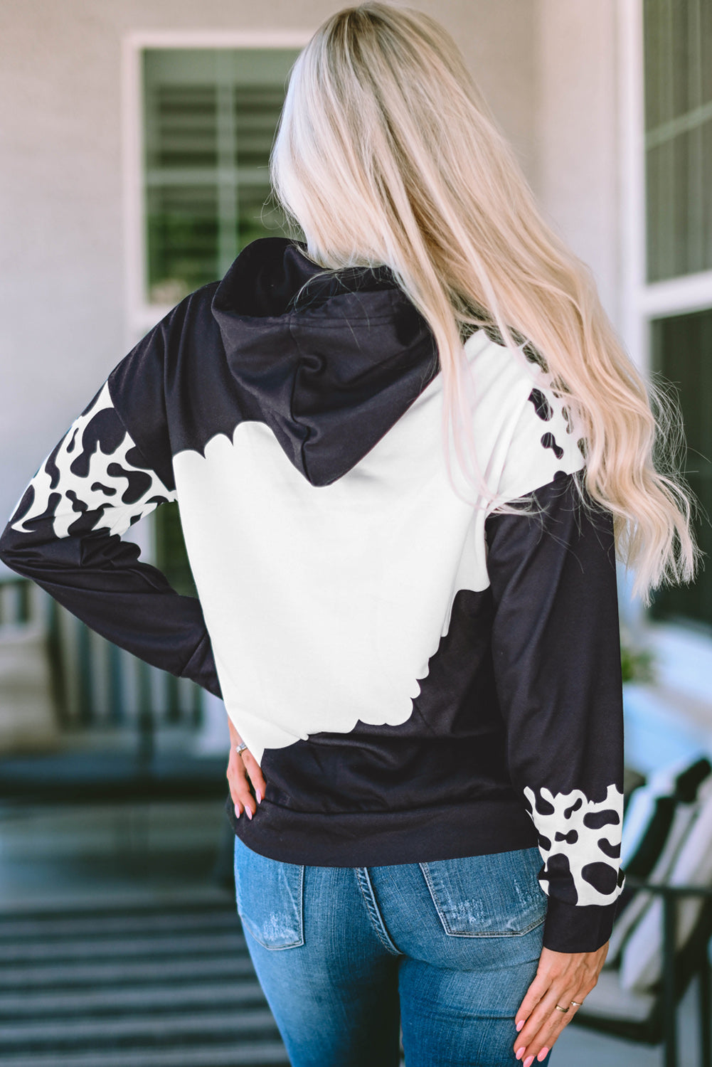 A person with long blonde hair is seen from the back wearing a Black Cow Tie Dye Print Pocketed Drawstring Pullover Hoodie and blue jeans. They are standing in an indoor space with a window and furniture in the background.