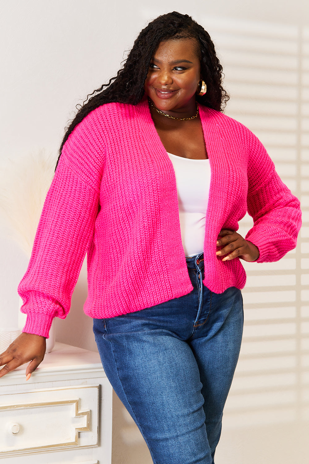 A woman wearing the Woven Right Rib-Knit Open Front Drop Shoulder Cardigan in bright pink over a white top and blue jeans stands indoors, looking to the side.