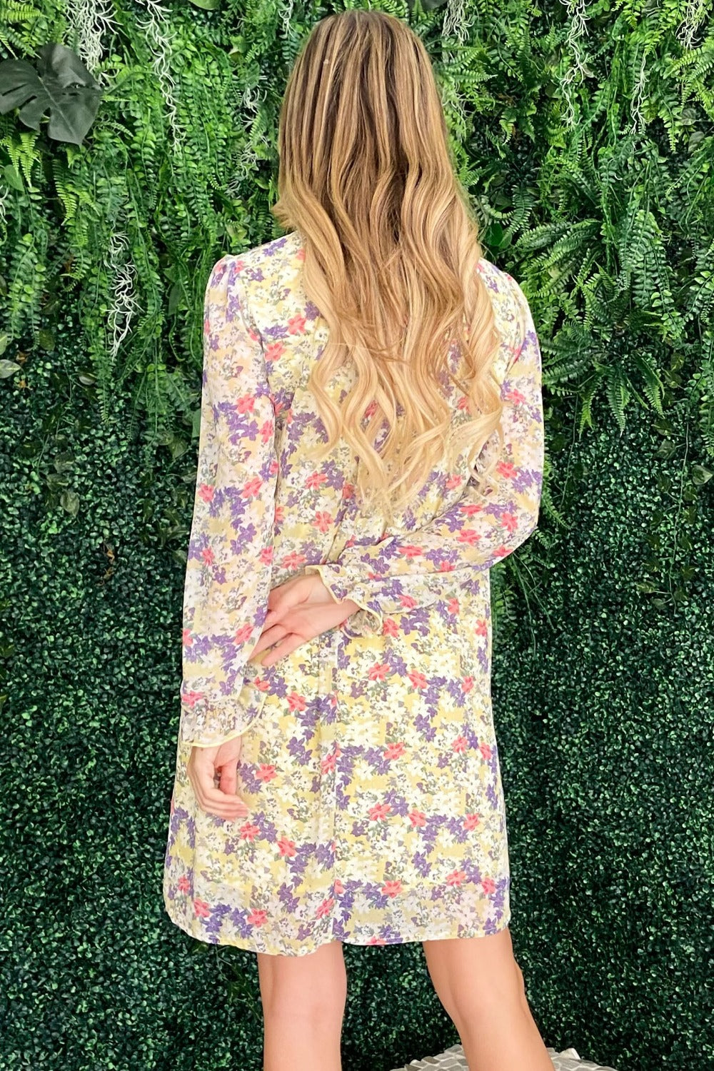 A woman stands in front of a leafy green background wearing the And The Why Floral Mock Neck Flounce Sleeve Dress, smiling and looking to the side.