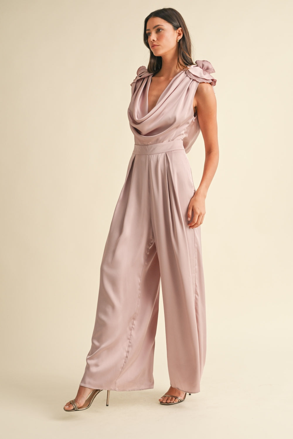 A woman stands against a plain background wearing the MABLE 3D Floral Applique Deep Cowl Neck Jumpsuit, which features an open back and wide-leg pants, paired with open-toed heels.