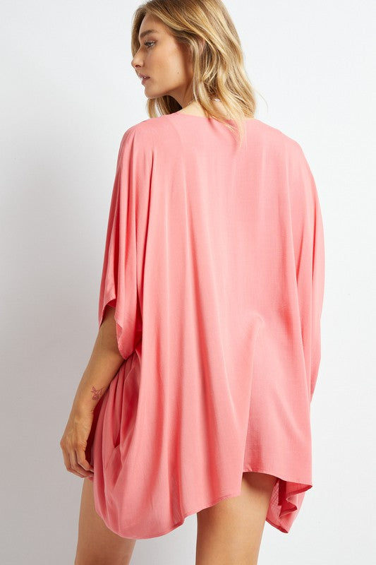 A woman poses against a simple backdrop, gracefully wearing a wardrobe essential: the Favorite Solid Kimono Cardigan in pink. Complementing her white tank top and denim shorts, this casual layer enhances her relaxed style with an elegant touch.