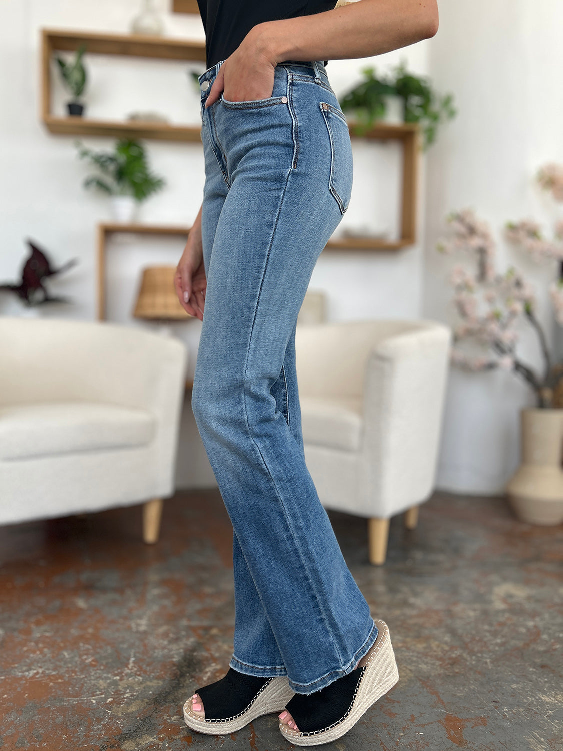 Person wearing Judy Blue Full Size Mid-Rise Waist Straight Jeans and a black top, standing in a room with white chairs, potted plants, and wooden shelves. Only the lower half of the body and part of the arms are visible.