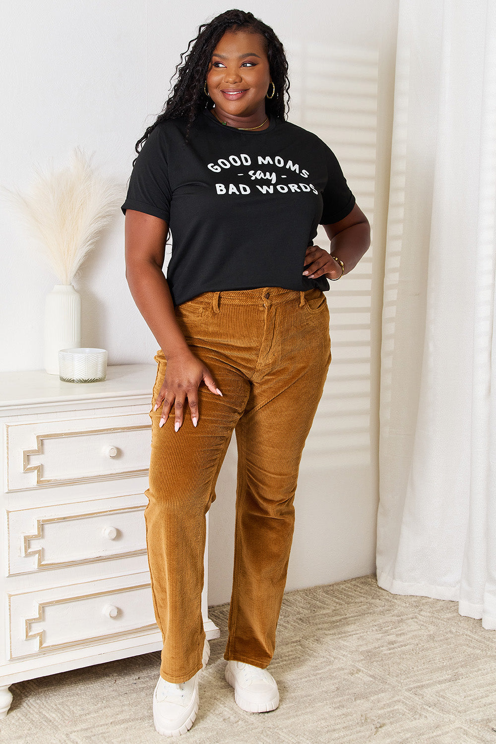 A woman stands in a room with a white wall, where light filters through the blinds, subtly showcasing her rebellious side while wearing the Simply Love GOOD MOMS SAY BAD WORDS Graphic Tee.