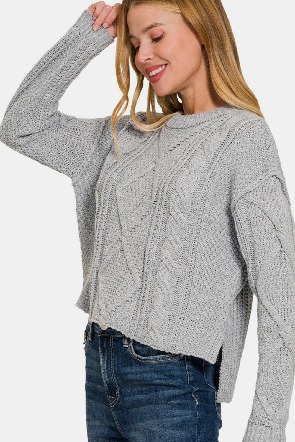 A person wearing a Zenana Cropped High Low Cable Sweater with Side Slits in light gray, paired with blue jeans, stands with their hand touching their head, smiling.