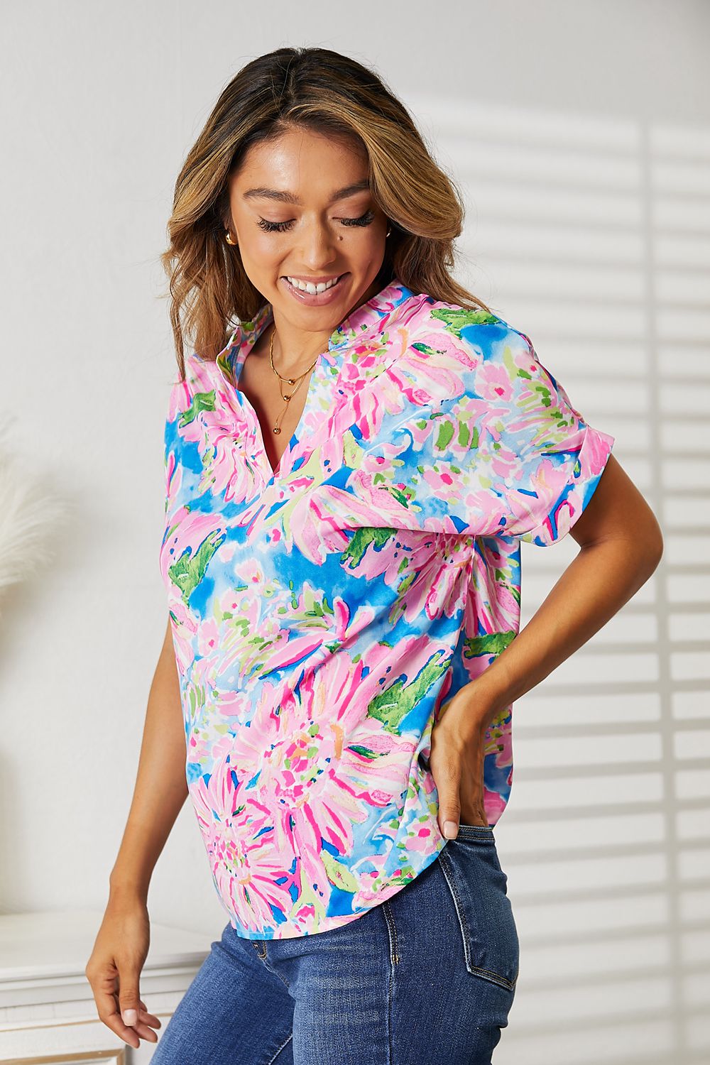 A woman stands indoors smiling, wearing a Double Take Floral Notched Neck Short Sleeve Top made of 100% polyester and blue jeans.