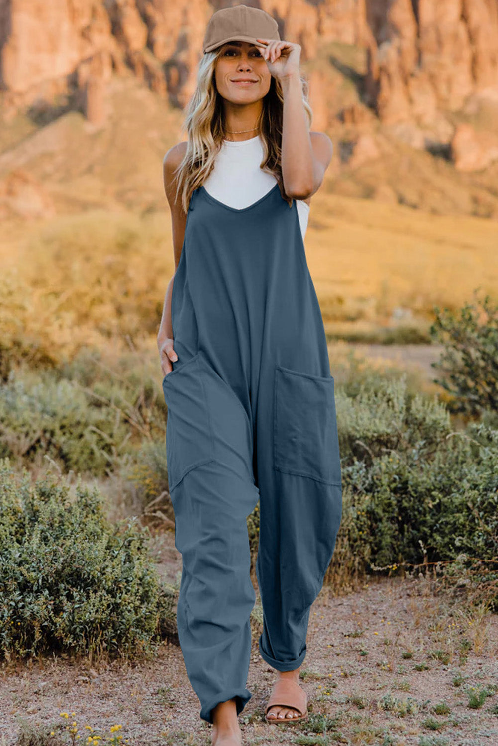 Person wearing a Double Take Full Size V-Neck Sleeveless Jumpsuit with Pockets and sandals, walking outdoors on a dirt trail with bushes in the background.