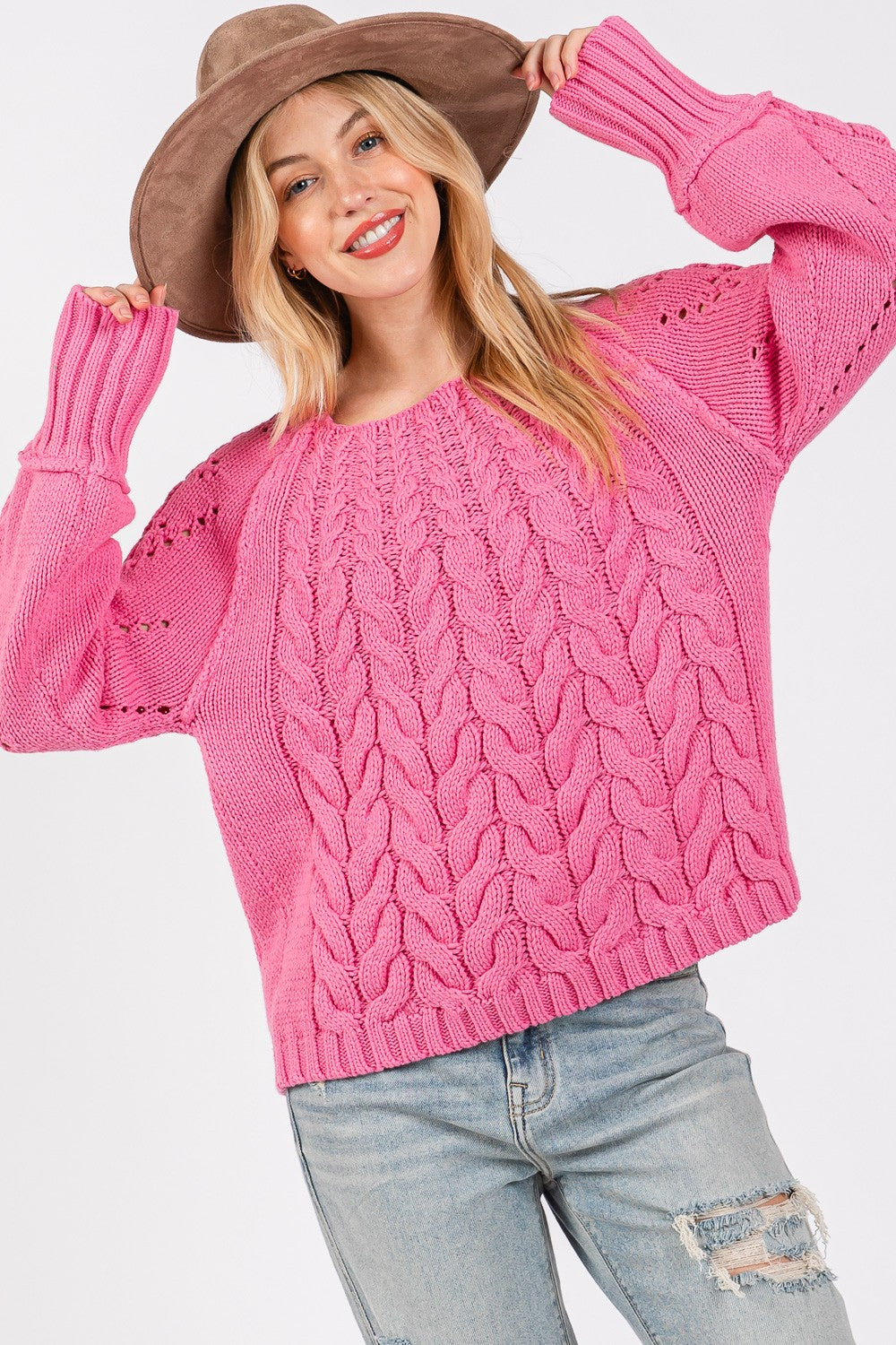 A person wearing a pink SAGE + FIG Cable-Knit Long Sleeve Sweater, light-wash ripped jeans, and a wide-brimmed hat poses with hands on hips.