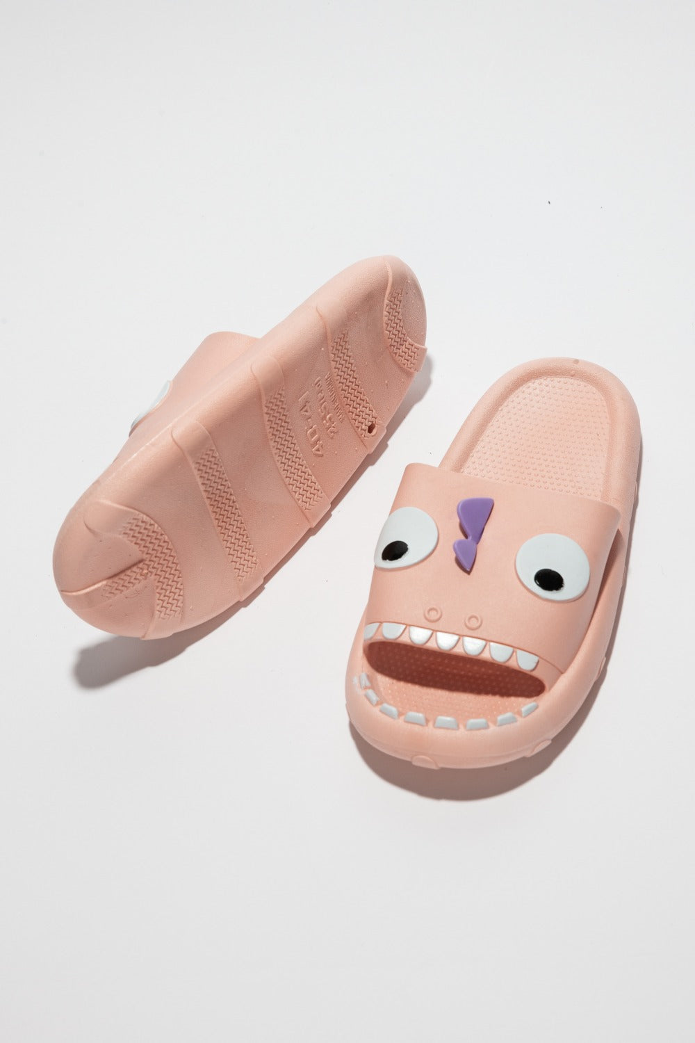 The NOOK JOI Monster Pillow Cloud Slides Non-Slip Slipper features cartoonish monster faces with large eyes, a purple nose, and an open mouth with teeth. Made of durable EVA material, these fun pink cloud slippers also boast an anti-slip design for added safety.