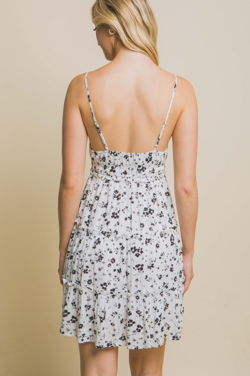 A woman wearing the Love Tree Floral Tiered Cami Mini Dress stands against a beige background, with one hand on her hip and a neutral expression.