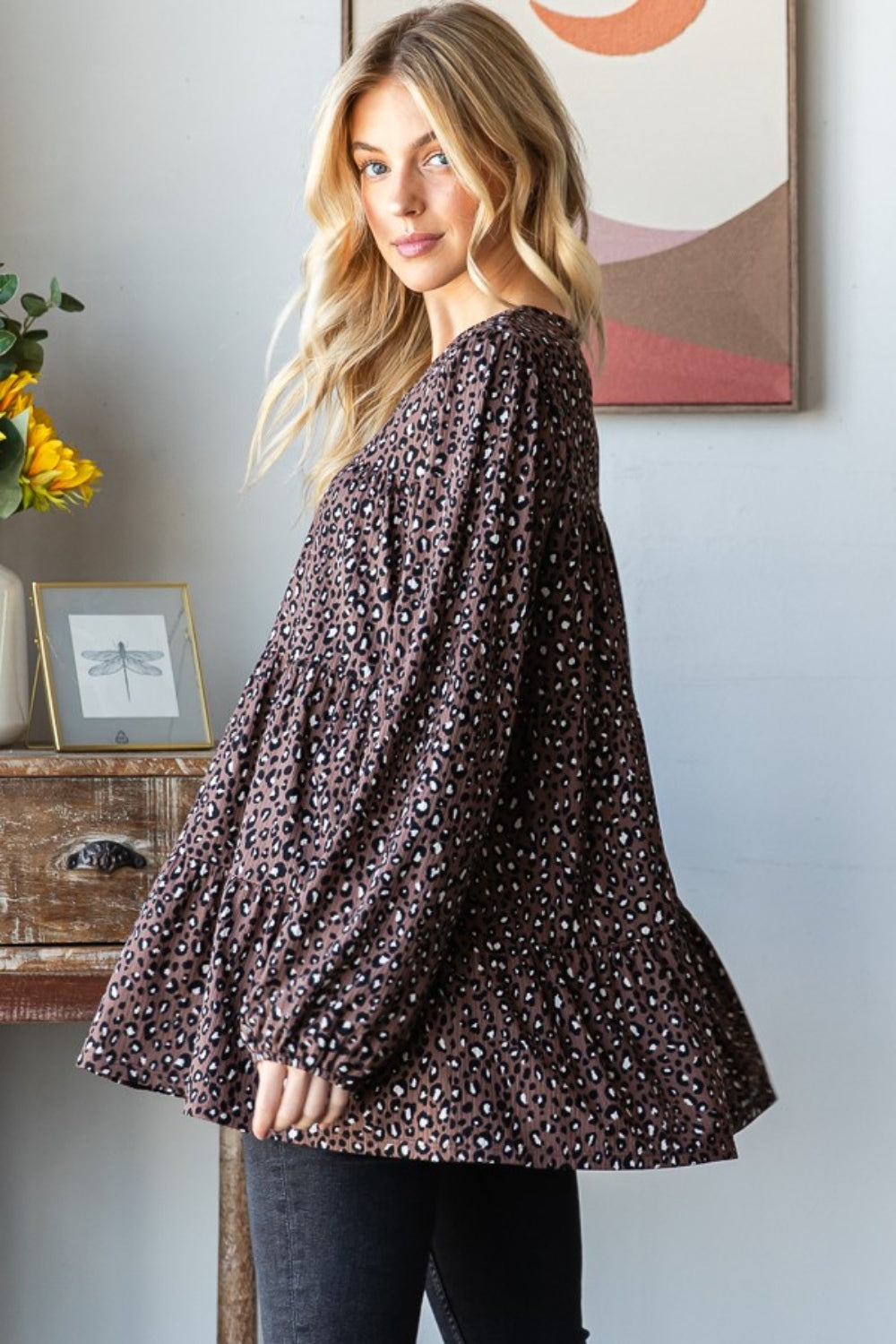 A woman wearing a Heimish Leopard Long Sleeve Tiered Blouse and black pants stands by a window indoors, looking at the camera—embodying a chic and feminine wardrobe.