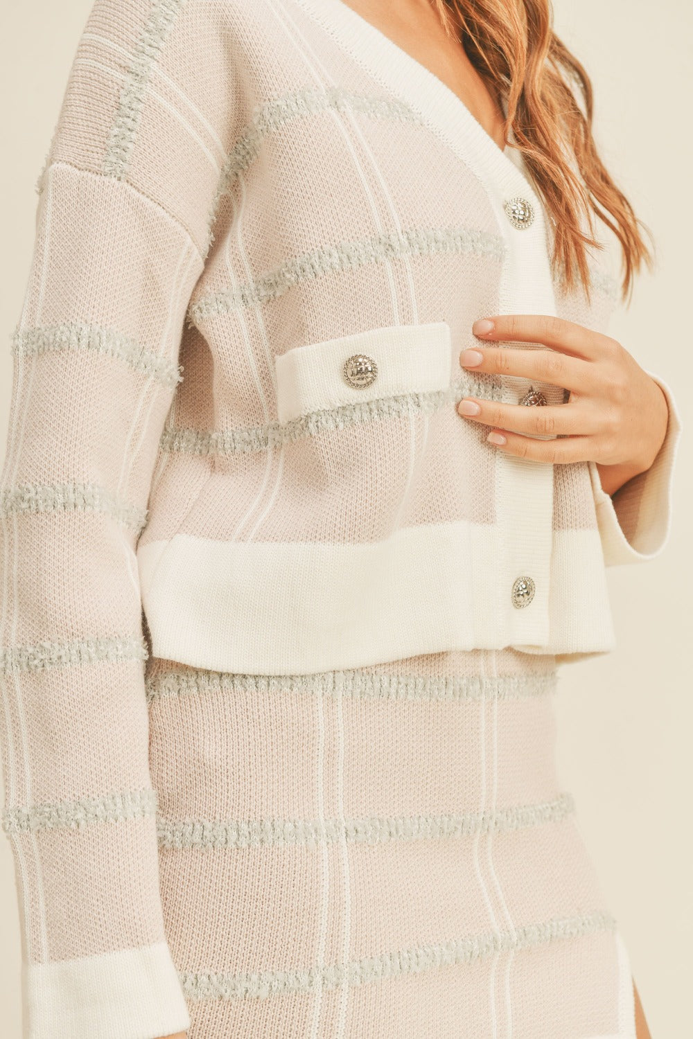 A woman stands wearing the MABLE Plaid Button Down Sweater Cardigan and Mini Skirt Set. The light-colored plaid cardigan features large buttons, perfectly matching the mini skirt. She has wavy hair and is touching her hip with one hand while looking at the camera.