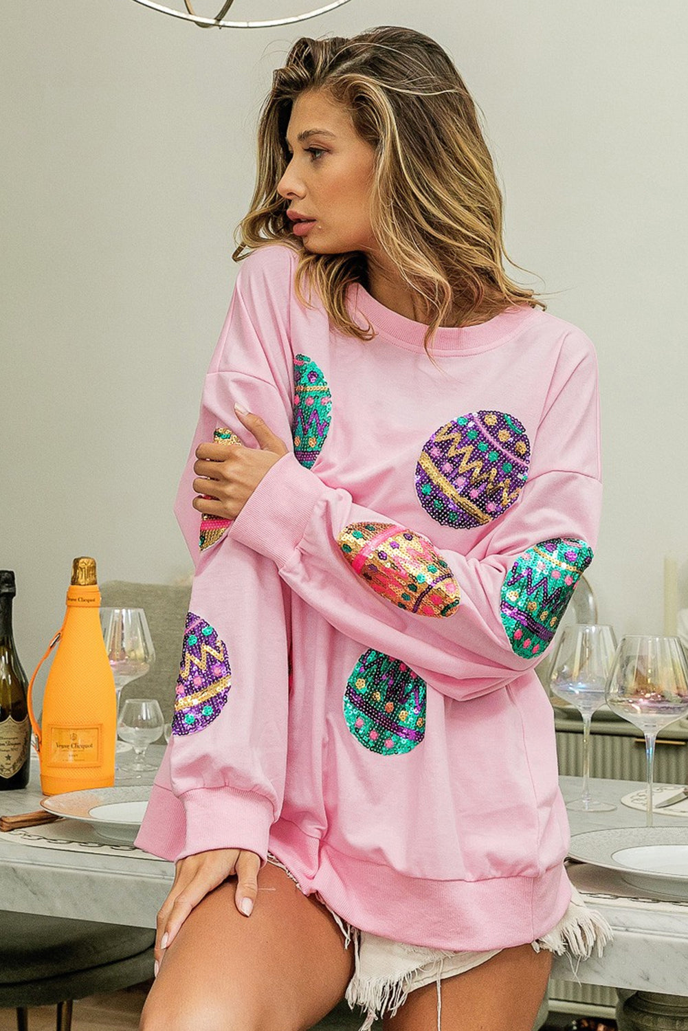 A woman with long, wavy hair wears a Pink Sequined Easter Egg Drop Shoulder Oversized Sweatshirt featuring colorful, patterned circles. She is seated in a dining area with a table set with glasses, plates, and two bottles, one of which is orange.
