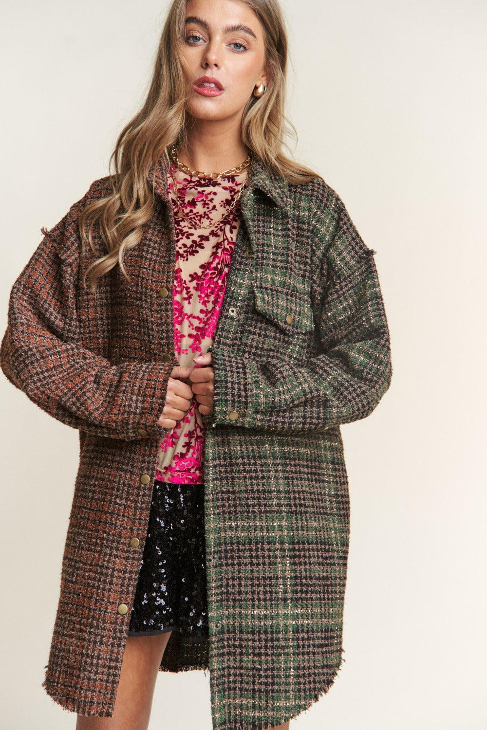 A woman wearing an eclectic mix poses against a plain background, showcasing an ideal fall wardrobe ensemble. She dons a mixed-patterned oversized jacket over a red floral sheer top and black sequined shorts. The J.NNA Snap Down Tweed Plaid Contrast Longline Shacket adds texture and depth to her look.