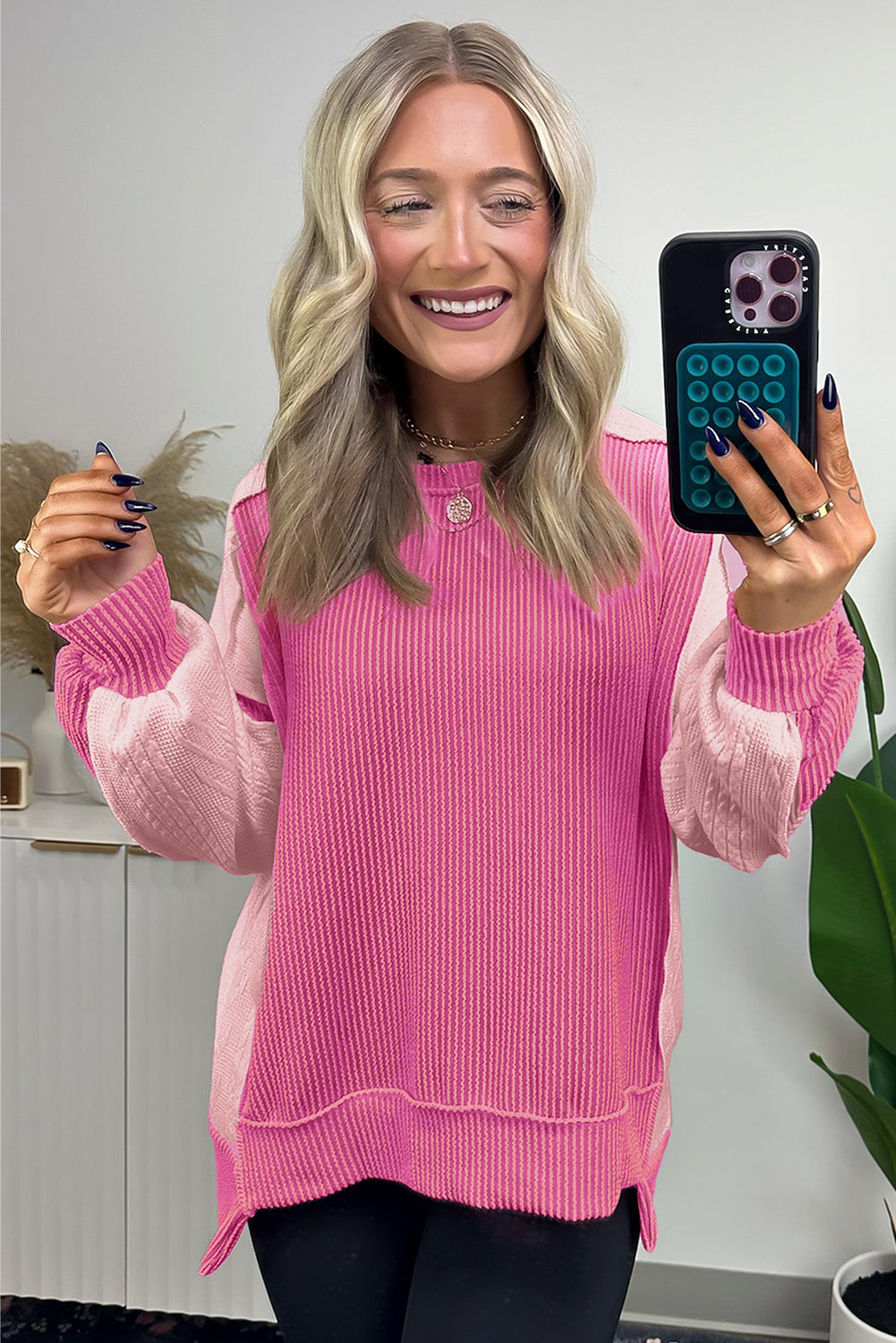 A person with long, wavy blonde hair is taking a selfie in a mirror, wearing the Bonbon Cable Knit Colorblock Exposed Seam Sweatshirt. A plant and decor items are visible in the background.