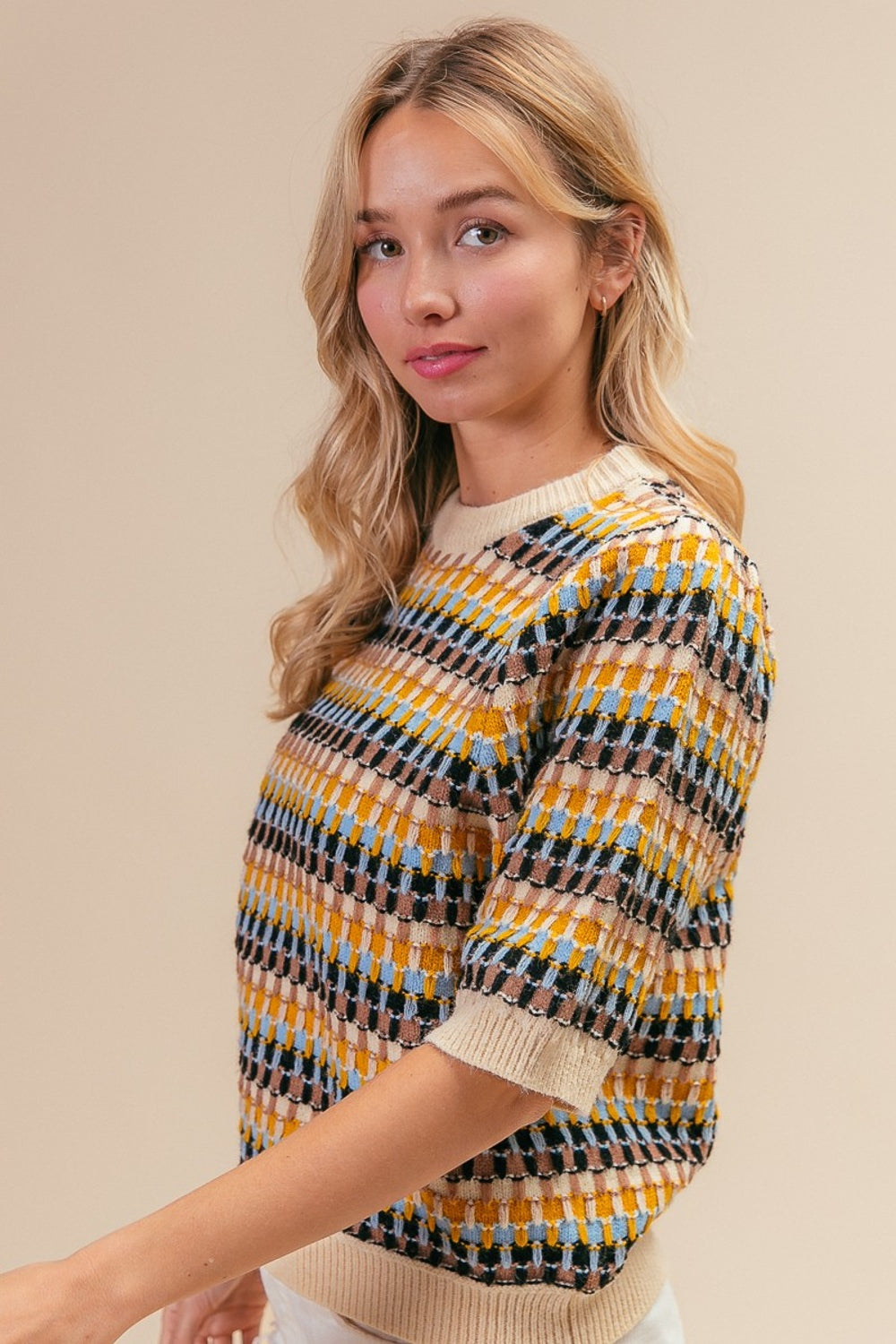 A woman with long blonde hair wearing a BiBi Multi Color Half Sleeve Sweater and white distressed shorts poses with one hand behind her head against a plain beige background.